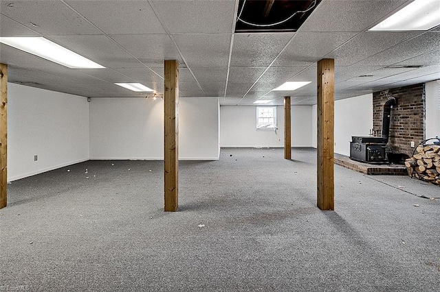 basement featuring a drop ceiling, carpet floors, baseboards, and a wood stove