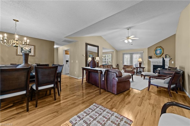 living room with light wood-type flooring, ceiling fan with notable chandelier, vaulted ceiling, a fireplace, and a textured ceiling