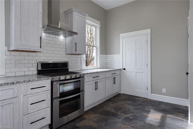 kitchen featuring light stone counters, backsplash, wall chimney range hood, range with two ovens, and white cabinets