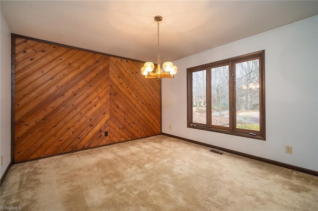 carpeted empty room featuring an inviting chandelier, baseboards, wooden walls, and visible vents