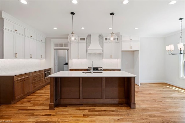 kitchen featuring light hardwood / wood-style flooring, a kitchen island with sink, hanging light fixtures, stainless steel microwave, and custom range hood