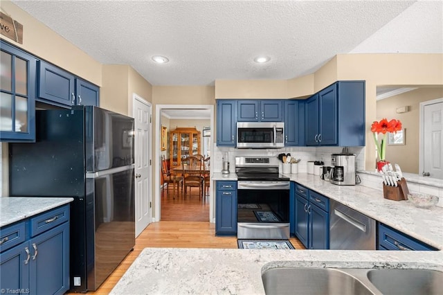 kitchen featuring blue cabinets, tasteful backsplash, stainless steel appliances, light wood-style floors, and glass insert cabinets