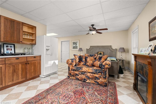 living area featuring light floors, baseboards, a ceiling fan, a drop ceiling, and a stone fireplace