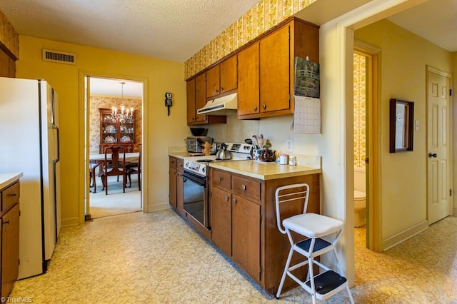 kitchen with an inviting chandelier, freestanding refrigerator, range with electric cooktop, under cabinet range hood, and brown cabinets