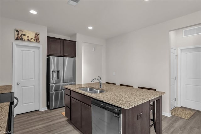 kitchen featuring appliances with stainless steel finishes, a kitchen island with sink, a sink, dark brown cabinetry, and light wood-type flooring