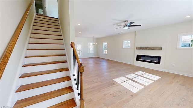 stairway with ceiling fan with notable chandelier, a glass covered fireplace, wood finished floors, recessed lighting, and baseboards