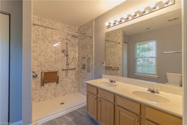 bathroom featuring double vanity, toilet, a tile shower, and a textured ceiling