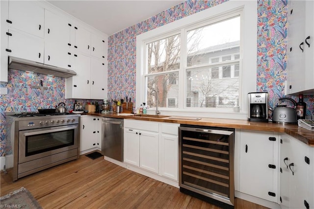 kitchen featuring wine cooler, under cabinet range hood, stainless steel appliances, wooden counters, and wallpapered walls