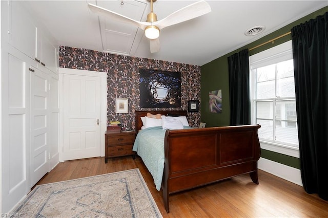 bedroom with ceiling fan, wood finished floors, visible vents, baseboards, and wallpapered walls