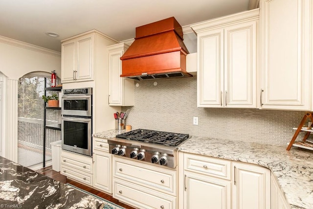 kitchen featuring stainless steel appliances, premium range hood, light stone counters, and cream cabinetry