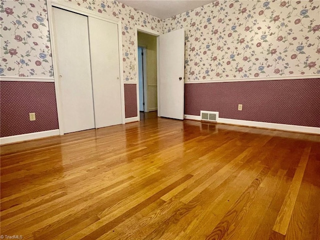 unfurnished bedroom with a wainscoted wall, a closet, visible vents, wood finished floors, and wallpapered walls