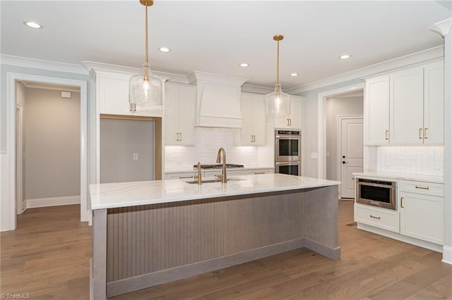 kitchen with double oven, hanging light fixtures, an island with sink, and premium range hood