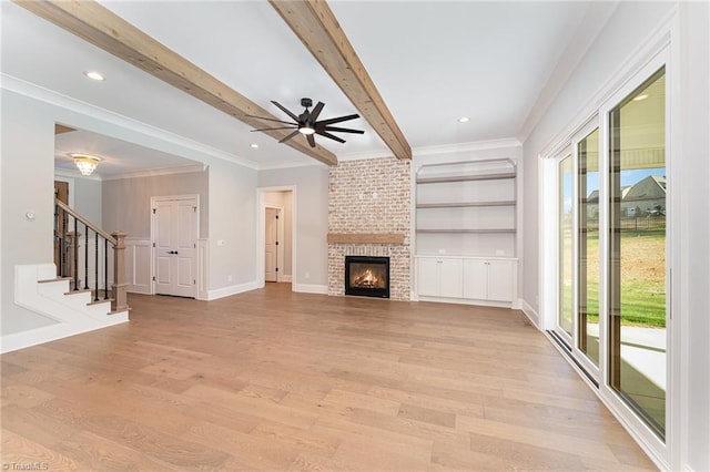 unfurnished living room featuring beamed ceiling, ceiling fan, crown molding, a brick fireplace, and light wood-type flooring