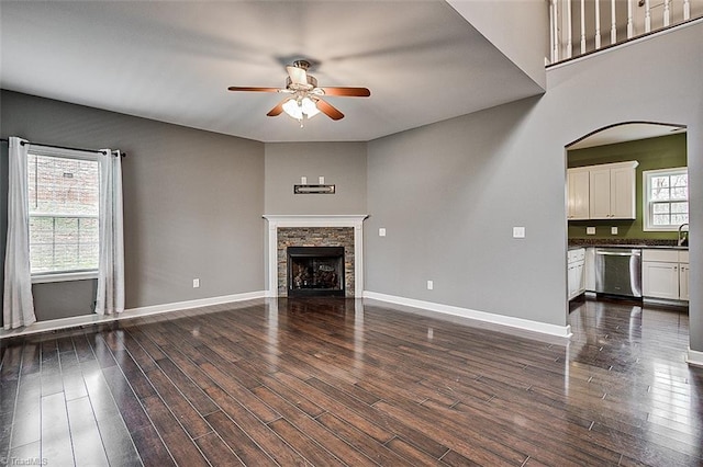 unfurnished living room with dark wood finished floors, a stone fireplace, a ceiling fan, and baseboards