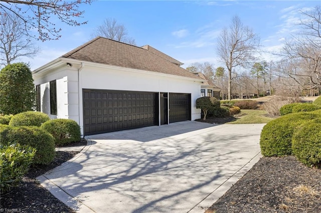 view of side of home with an attached garage, a shingled roof, concrete driveway, and stucco siding