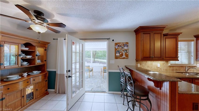kitchen featuring dark stone counters, brown cabinetry, ceiling fan, backsplash, and a textured ceiling