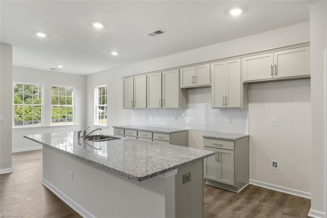 kitchen featuring sink, gray cabinets, a kitchen island with sink, light stone countertops, and decorative backsplash