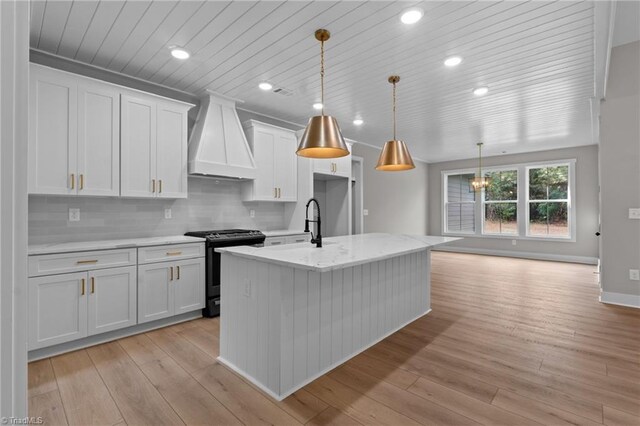 kitchen featuring range with gas cooktop, decorative light fixtures, an island with sink, white cabinets, and custom exhaust hood