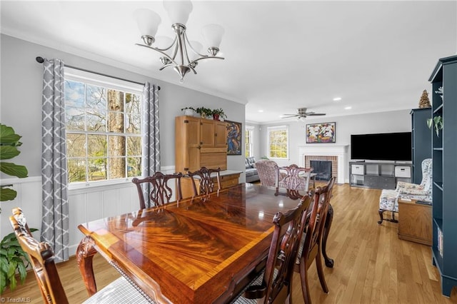 dining area featuring light wood-type flooring, ornamental molding, wainscoting, ceiling fan with notable chandelier, and a fireplace