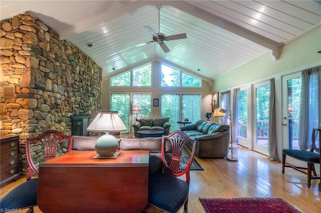 living room featuring light hardwood / wood-style floors, beamed ceiling, high vaulted ceiling, a stone fireplace, and ceiling fan