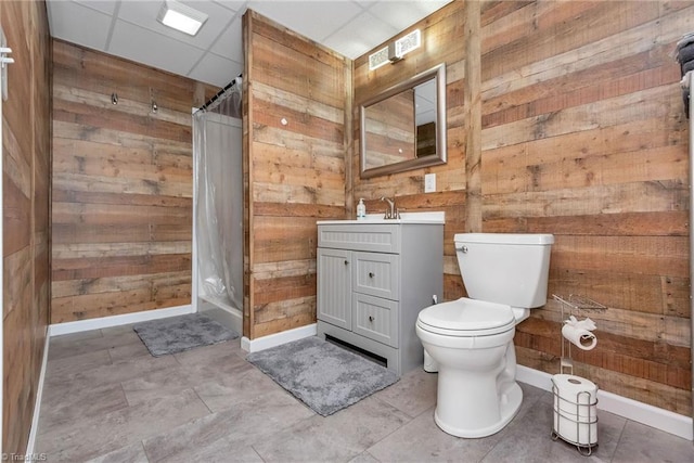 bathroom with a drop ceiling, walk in shower, toilet, wooden walls, and vanity