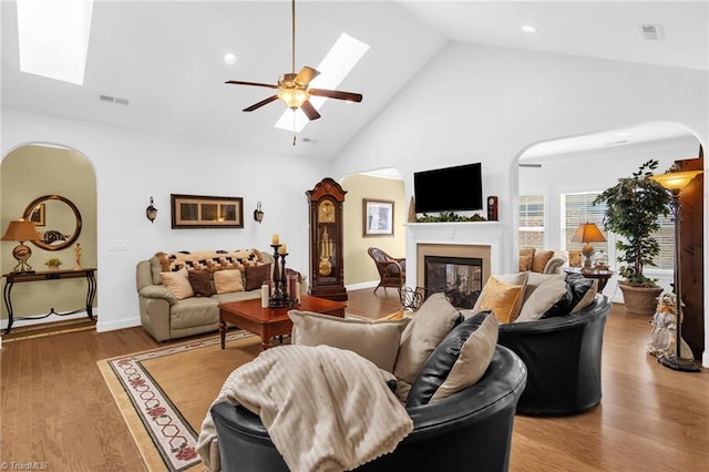 living room with arched walkways, a skylight, high vaulted ceiling, a glass covered fireplace, and a ceiling fan