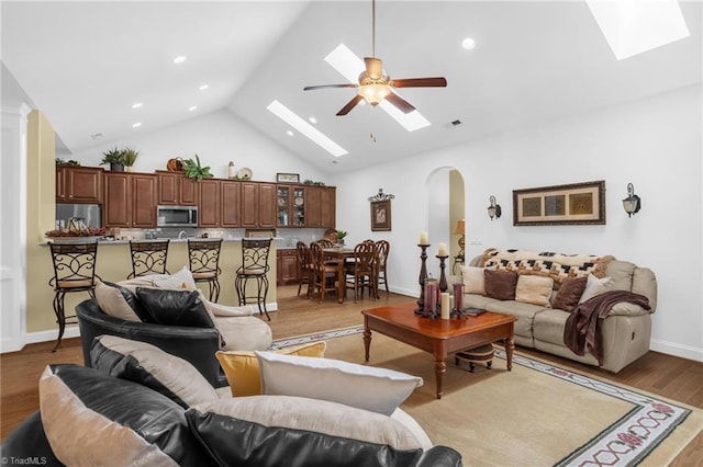 living room featuring a skylight, arched walkways, ceiling fan, high vaulted ceiling, and light wood finished floors
