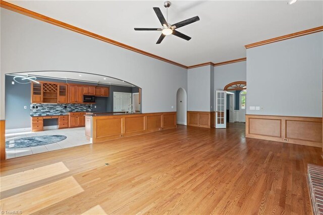 unfurnished living room featuring a ceiling fan, arched walkways, a wainscoted wall, ornamental molding, and light wood-style flooring
