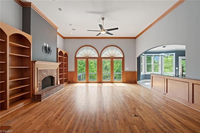 unfurnished living room with a ceiling fan, arched walkways, a brick fireplace, wood finished floors, and ornamental molding