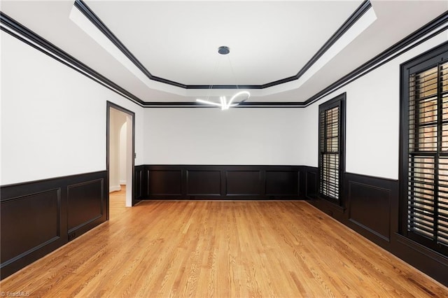 empty room featuring crown molding, wainscoting, a chandelier, a tray ceiling, and light wood-style flooring