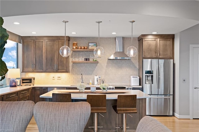 kitchen with hanging light fixtures, wall chimney exhaust hood, decorative backsplash, light hardwood / wood-style flooring, and stainless steel fridge