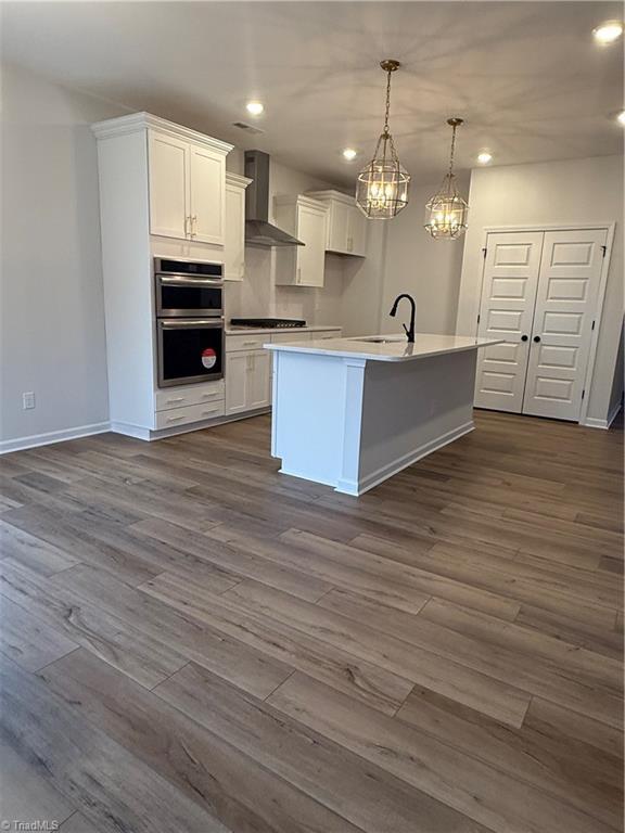 kitchen featuring a kitchen island with sink, double oven, white cabinetry, decorative light fixtures, and wall chimney exhaust hood