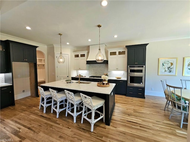 kitchen with custom exhaust hood, sink, a center island with sink, light hardwood / wood-style flooring, and white cabinetry