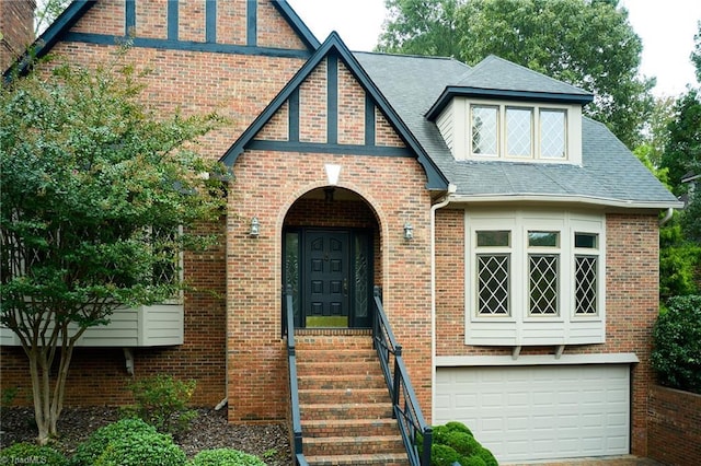 english style home featuring a garage, brick siding, driveway, and roof with shingles