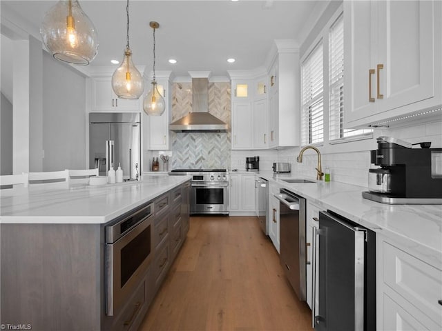 kitchen featuring tasteful backsplash, wall chimney range hood, built in appliances, white cabinets, and hanging light fixtures