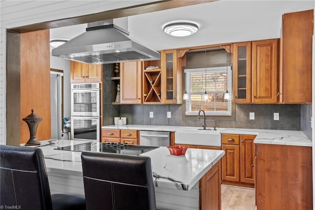 kitchen with decorative backsplash, appliances with stainless steel finishes, brown cabinets, wall chimney range hood, and a sink