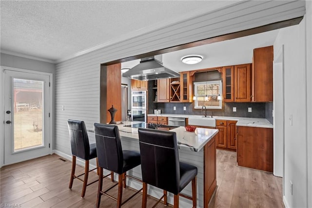 kitchen featuring wall chimney exhaust hood, appliances with stainless steel finishes, brown cabinets, a kitchen bar, and a sink