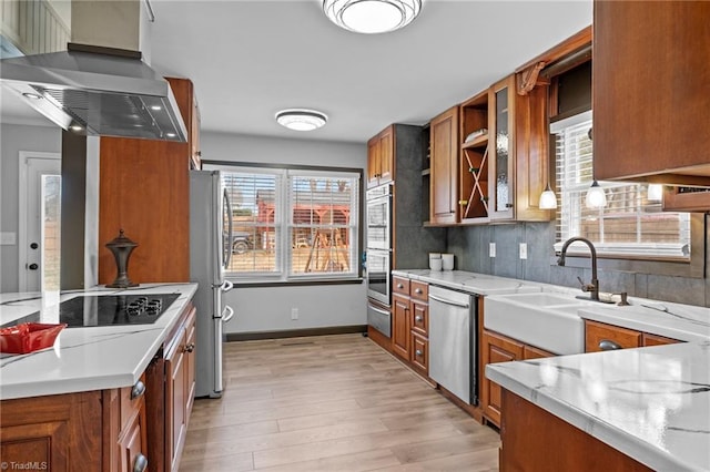 kitchen featuring a warming drawer, appliances with stainless steel finishes, brown cabinetry, light wood-type flooring, and wall chimney exhaust hood