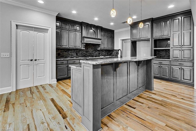 kitchen featuring hanging light fixtures, light stone counters, crown molding, a center island with sink, and light wood-type flooring