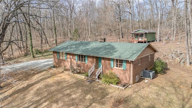 ranch-style house featuring brick siding, driveway, a view of trees, and central air condition unit