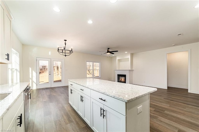 kitchen featuring ceiling fan with notable chandelier, a warm lit fireplace, dark wood-style floors, a center island, and recessed lighting