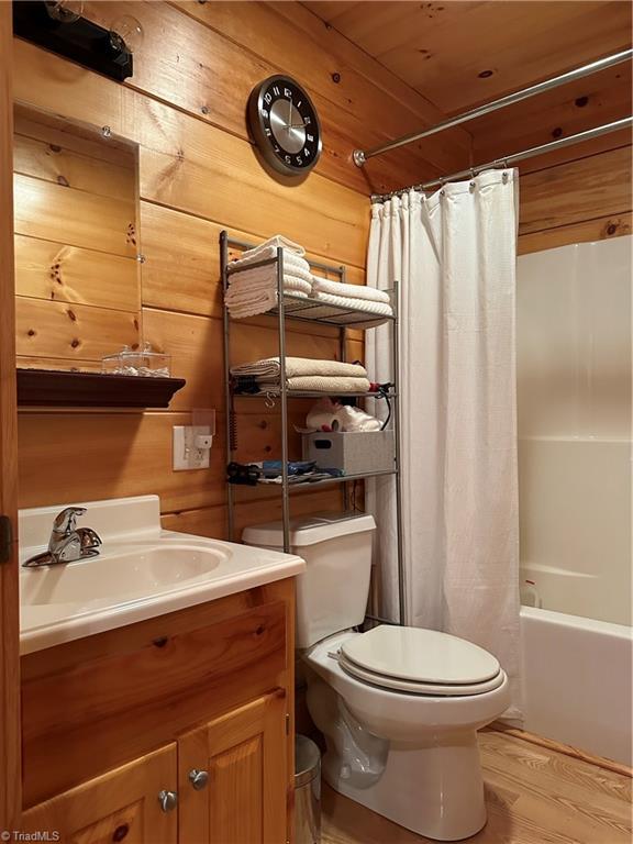 full bathroom with wood ceiling, vanity, wooden walls, shower / bathtub combination with curtain, and toilet