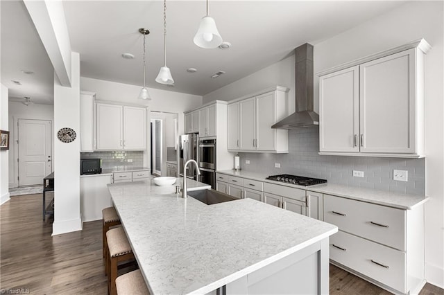 kitchen featuring wall chimney exhaust hood, a sink, appliances with stainless steel finishes, a kitchen bar, and an island with sink