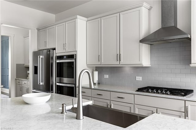kitchen featuring stainless steel appliances, wall chimney exhaust hood, decorative backsplash, a sink, and light stone counters