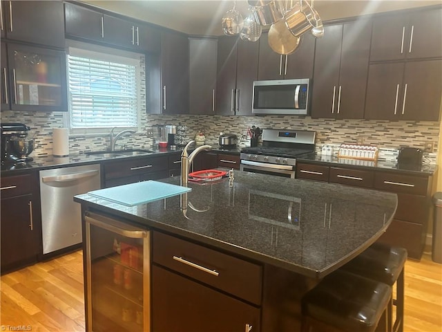 kitchen featuring beverage cooler, a center island with sink, light wood-style flooring, stainless steel appliances, and a sink