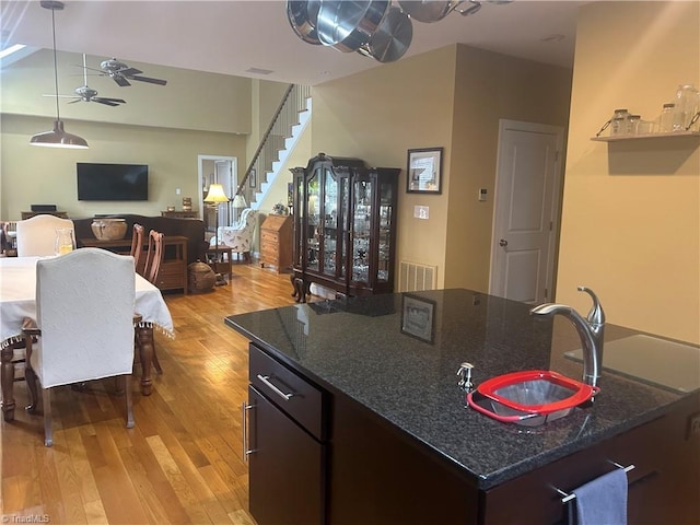 kitchen with light wood-style floors, a ceiling fan, a kitchen island with sink, a sink, and dark stone countertops