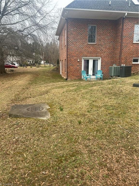 back of house featuring a shingled roof, cooling unit, brick siding, and a lawn