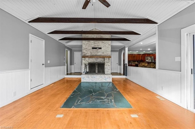 unfurnished living room featuring lofted ceiling with beams, ceiling fan, a fireplace, wood finished floors, and wainscoting