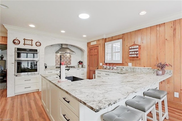 kitchen with wood walls, light wood-type flooring, stainless steel double oven, and wall chimney exhaust hood