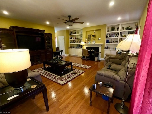 living area with a ceiling fan, built in shelves, wood finished floors, a fireplace, and recessed lighting
